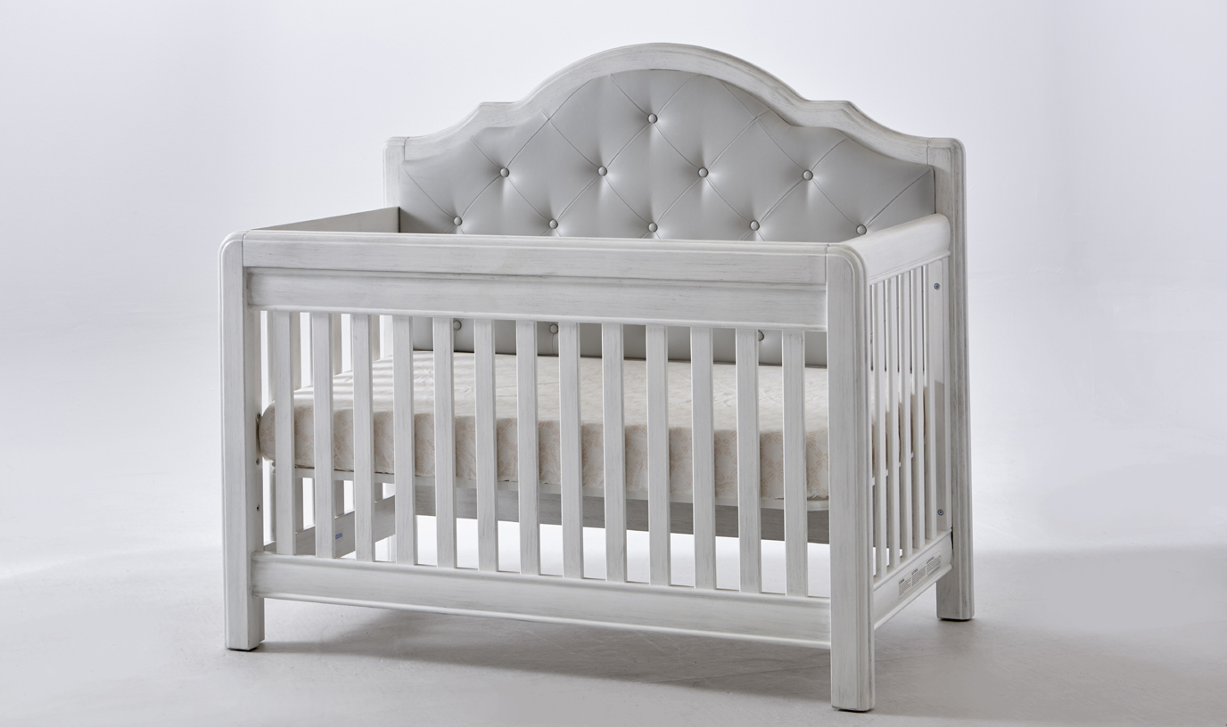 The <b>Cristallo Forever Crib</b> in Vintage White with a gray vinyl upholstered panel headboard. <br>Like the playful nature of light that bounces off a crystal and makes rainbows on the wall, the architectural shapes paired with the upholstered cushion of the Cristallo Forever Crib provide a playful combination of refined styling and chic design.  The Cristallo Forever Crib is an enchanting blend of classic lines paired with sweet touches of sophisticated luxury.  The strong, clean design of this crib is highlighted by the layered moldings that frame both the front and the headpiece.  These beautifully rounded layers provide texture and a nod to the elegance of classical architecture.  Set in the headpiece of the crib is a sumptuously tufted panel. Providing a distinct sense of playful beauty, this sweetly textured cushion is designed with both style and use in mind.  